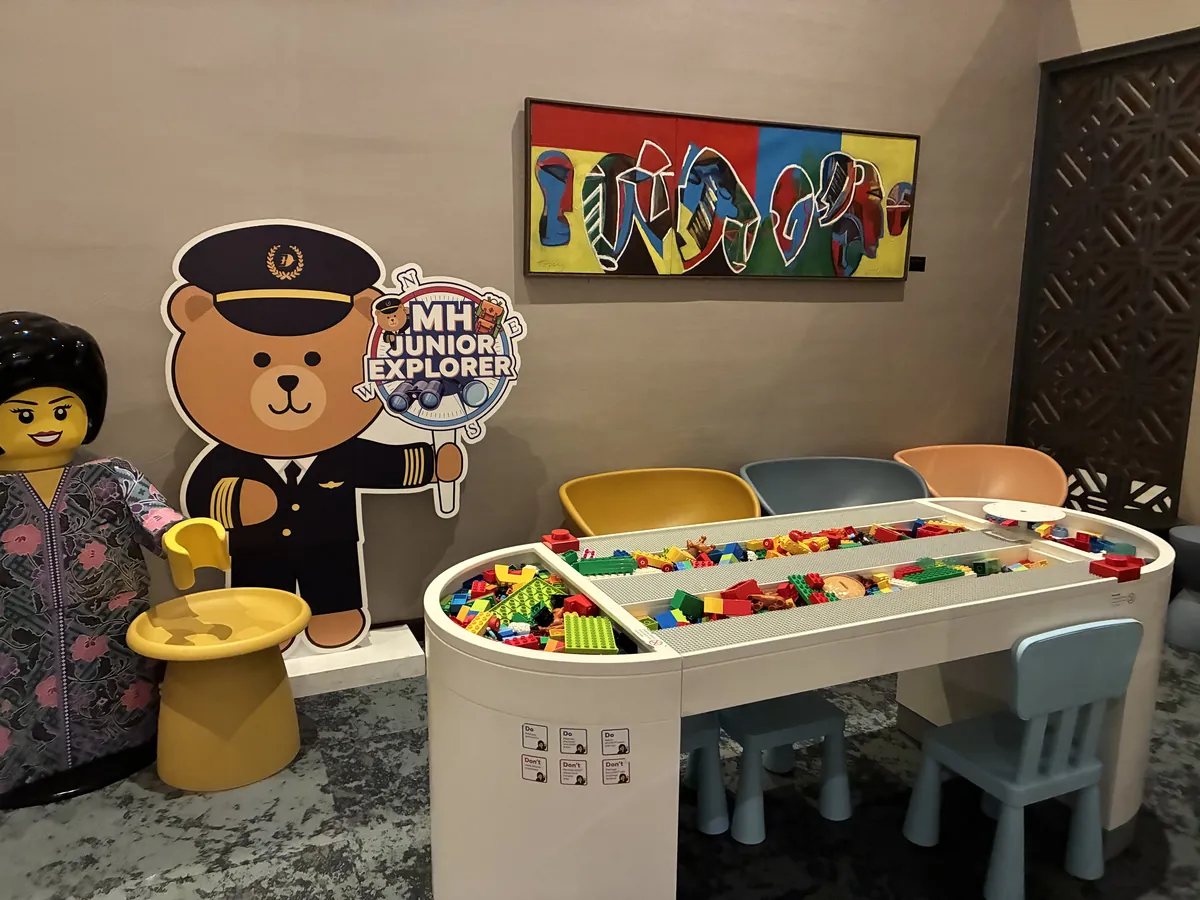 LEGOブロックがあふれるビルディングテーブルと、パイロット帽をかぶった「MH JUNIOR EXPLORER」のクマの看板、マレーシアの民族衣装を着た大きなLEGO人形