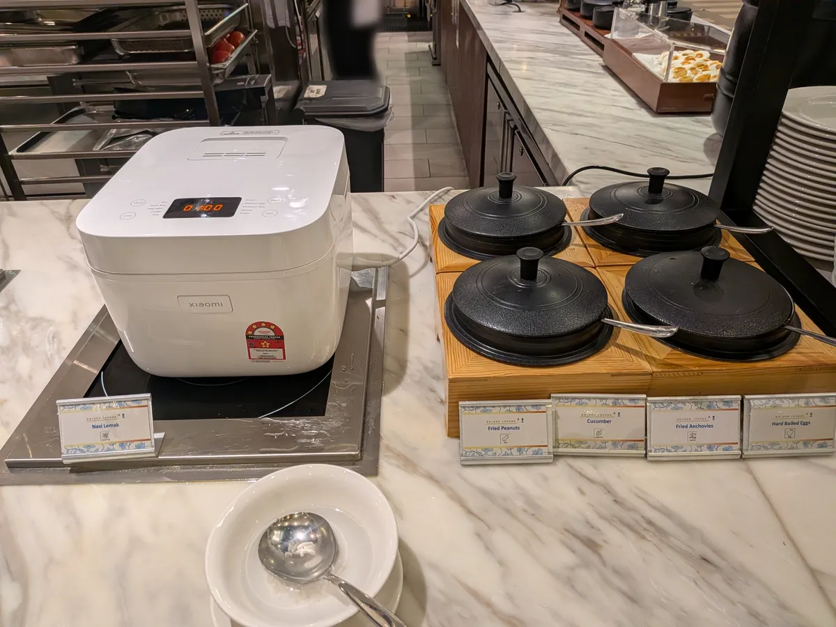 ナシレマッの炊飯器と、黒い蓋つきの器に入った4種のコンディメント（揚げピーナッツ、きゅうり、イカンビリス、ゆで卵）