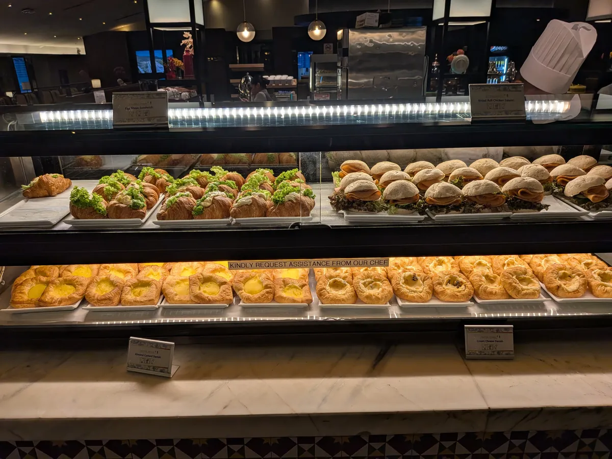 パンのショーケース。クロワッサンサンド、チャバタのハムサンド、デニッシュが並び、「KINDLY REQUEST ASSISTANCE FROM OUR CHEF」の表示とシェフ帽が見える