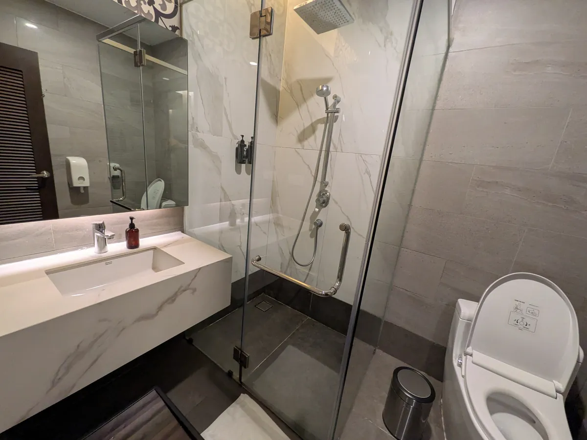 個室のシャワールーム。大理石調の洗面台とガラス張りのレインシャワー、トイレが同室に備わる