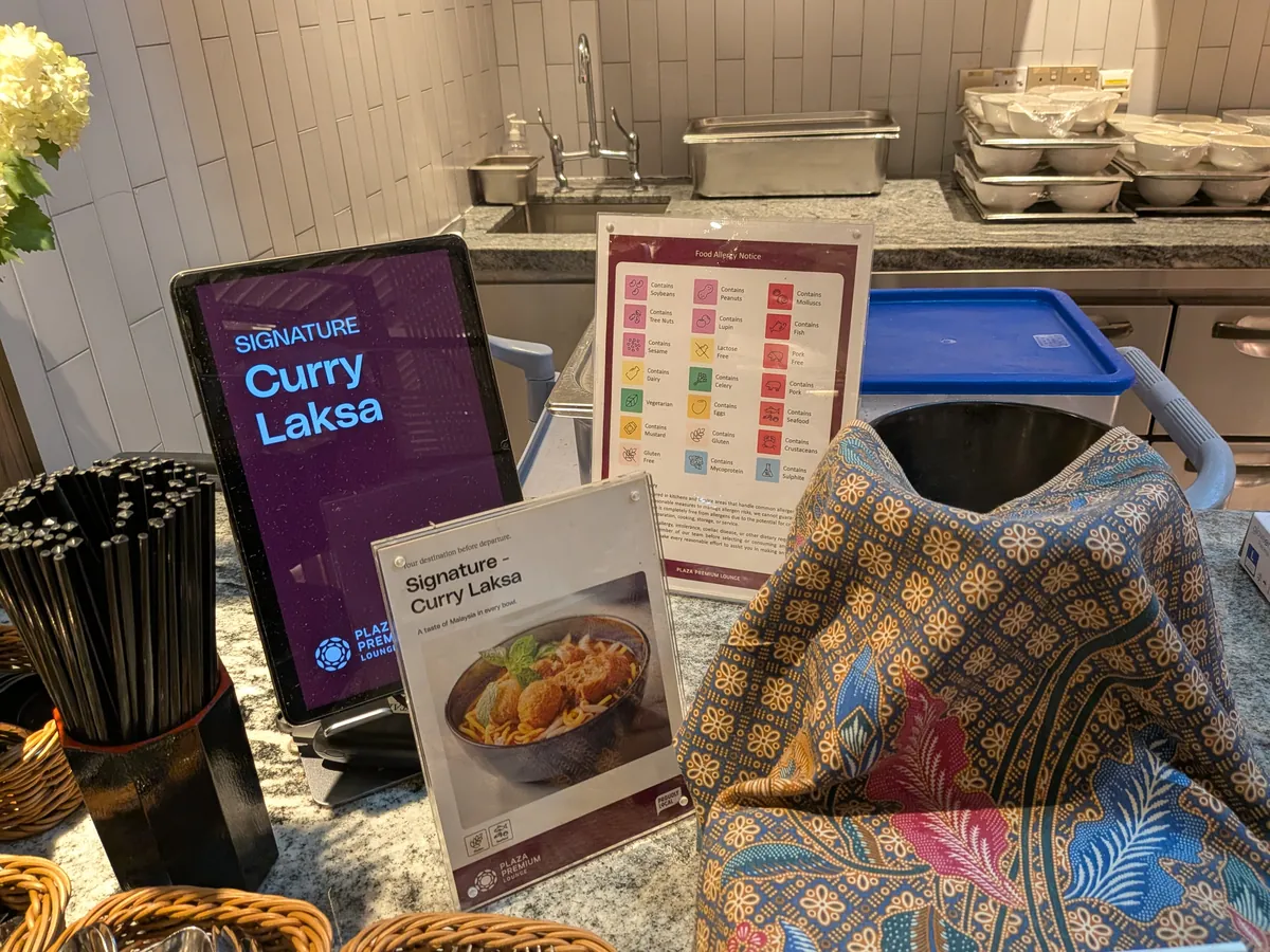 シグネチャーメニュー「Curry Laksa」のサイネージと写真付きカード。アレルゲン表示のボードとバティックの布を掛けた寸胴が並ぶヌードルステーション