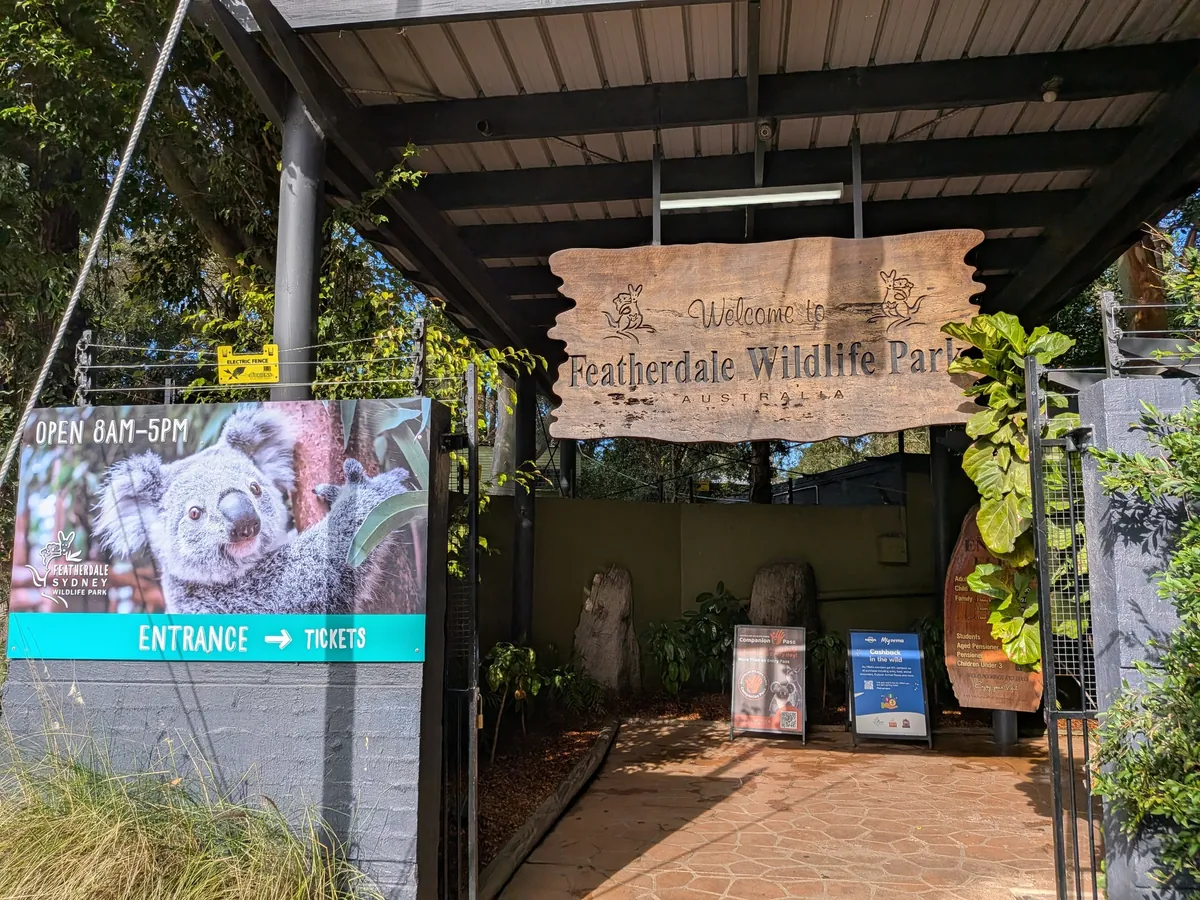 フェザーデール・ワイルドライフパークのウェルカムゲート。木の看板と、コアラの写真に「OPEN 8AM-5PM/ENTRANCE→TICKETS」と書かれた案内板が並ぶ