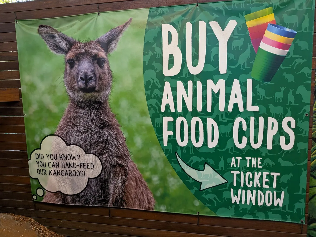「BUY ANIMAL FOOD CUPS AT THE TICKET WINDOW」と大書された園内のバナー。カンガルーの写真と色とりどりの餌カップのイラストが添えられている