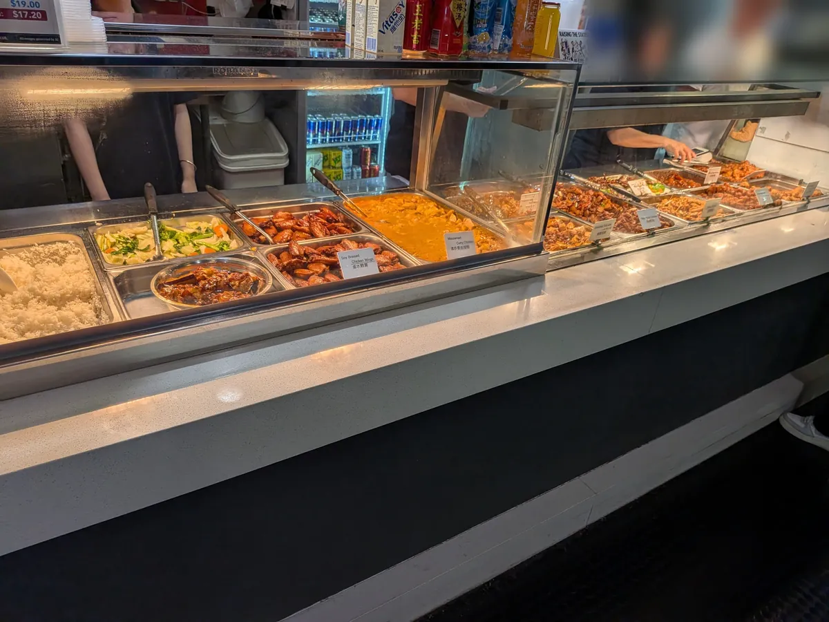 Little Asiaのショーケース。蒸し野菜や手羽の甘辛煮、カレー、マーボー豆腐など十数種類の惣菜がバットに並び、上に19.00豪ドルと17.20豪ドルの2段価格の値札が下がっている