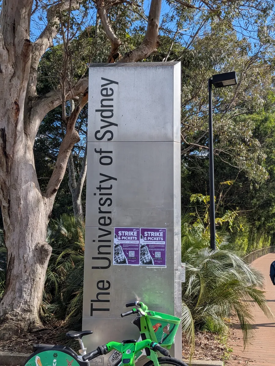 キャンパス入口に立つ「The University of Sydney」と縦書きされた石の看板柱。奥にユーカリの大木が茂る