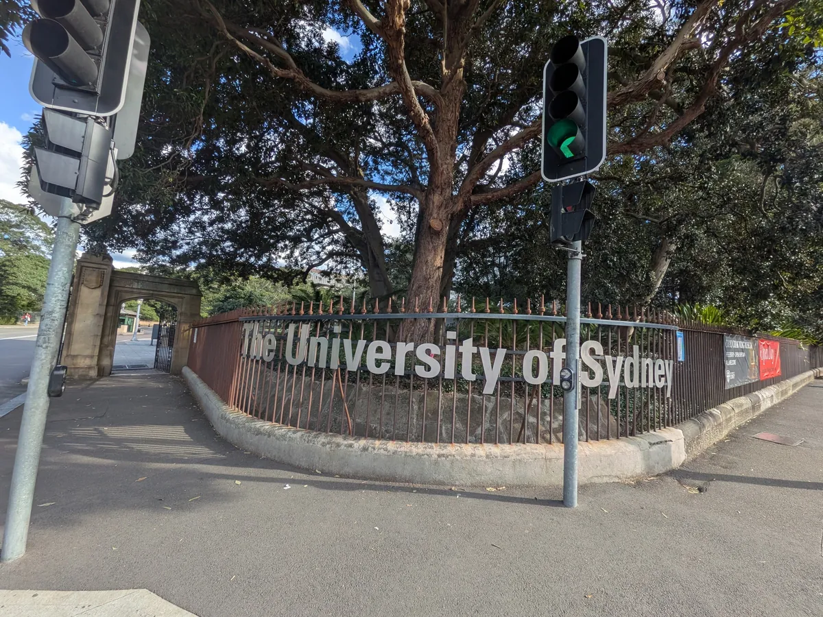 シドニー大学の門。鉄柵に「The University of Sydney」の切り文字が入り、奥に砂岩の建物と大きな木が見える
