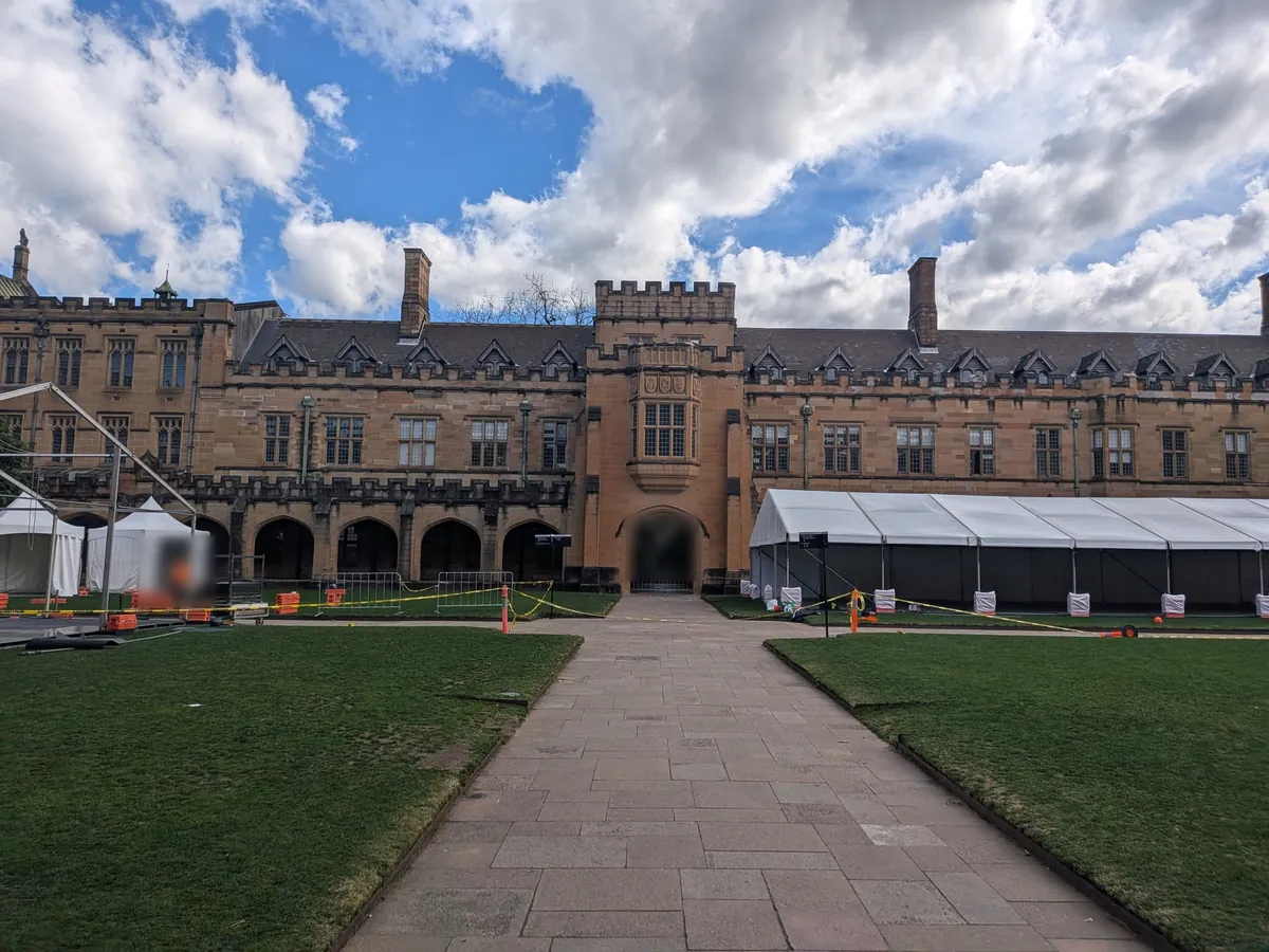 シドニー大学クアドラングルの中庭。行事の設営中で白い大型テントとオレンジのバリケードが並び、正面のアーチ門へ石畳の道が伸びている