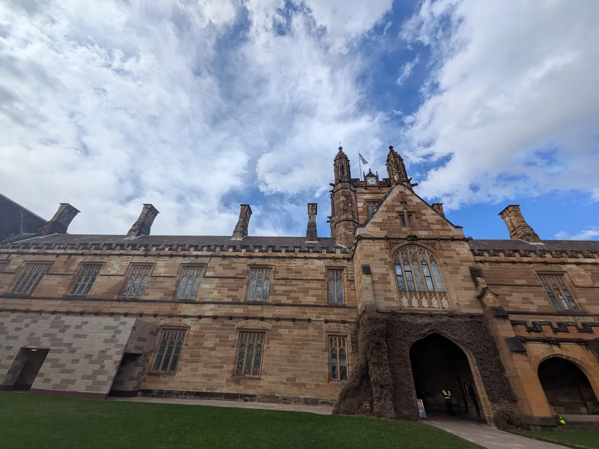 シドニー大学クアドラングルの回廊ファサード。砂岩のアーチと尖った切妻、狭間のある屋根が青空を背景に連なる