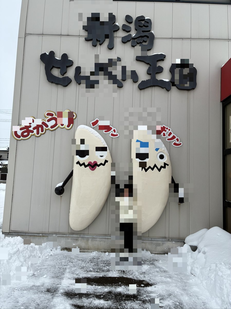 雪が積もる新潟せんべい王国の建物と、巨大なせんべいキャラクターオブジェの前でポーズをとるメイ