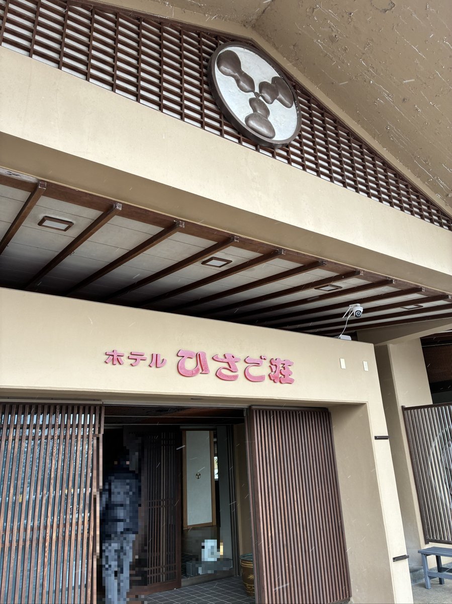 「ホテルひさご荘」の入り口外観。雪が舞い散る中、引き戸が開かれ、宿泊客を迎え入れている様子。上部には紋のようなデザインが見える。