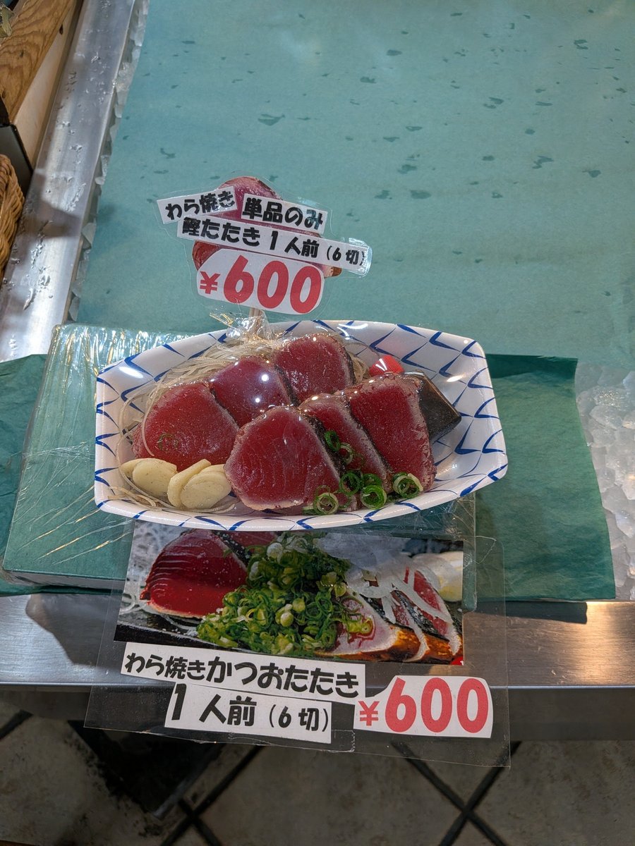 皿に盛り付けられた新鮮なカツオのたたきと、添えられたニンニクスライス、ネギ。手書きのポップには「わら焼きかつおたたき 1人前 (6切) ¥600」とある。