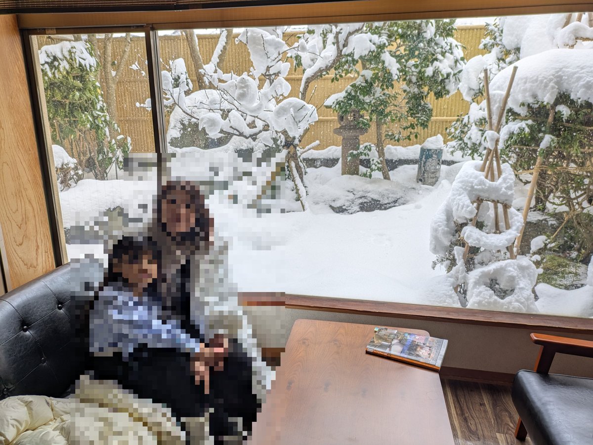 大きな窓から雪景色が広がる和風の部屋。窓の外には雪が深く積もった庭園が広がり、冬の風情を感じさせる。室内は暖かく快適な様子。
