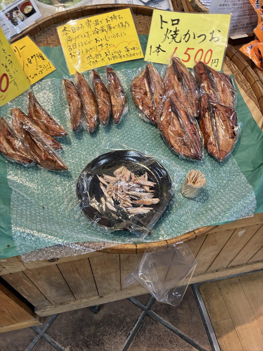 木製の台の上に、真空パックされた干物や小魚の加工品が複数並べられている。手書きのポップには「トロ焼かつお 1本 1500円」などの文字が見える。