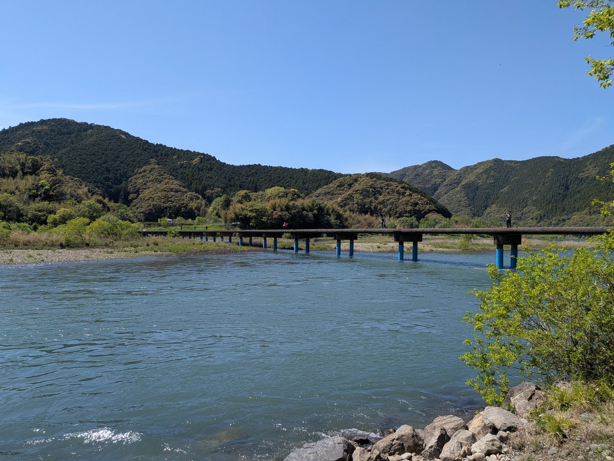 四万十川に架かる佐田沈下橋の全景。水色の橋脚が特徴的な橋が川面に長く伸び、両岸には豊かな緑の山々が連なる。青い空が広がる穏やかな風景。