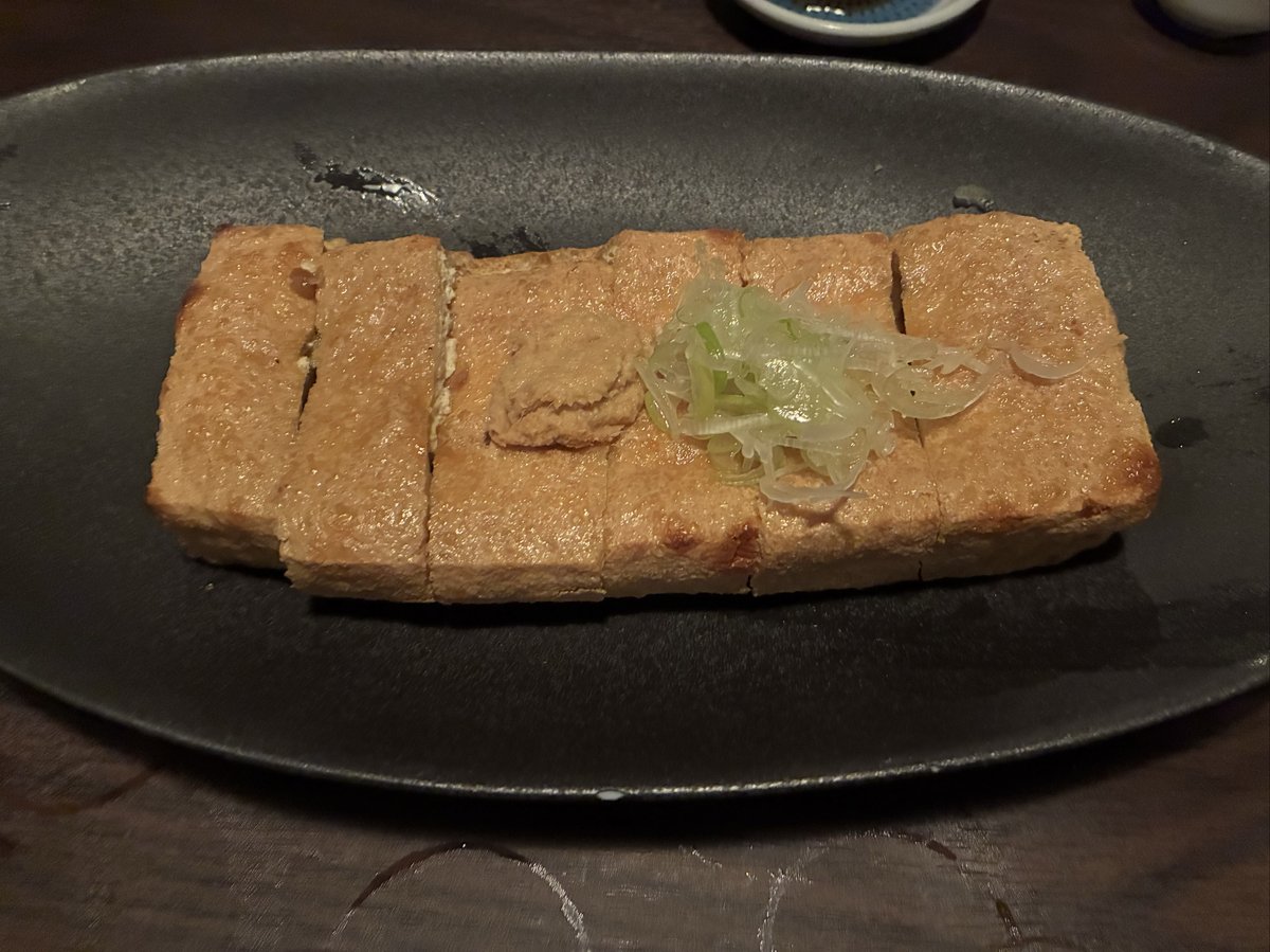 越後番屋酒場の厚揚げ焼き。黒い皿に乗った分厚い豆腐が香ばしく焼き上げられ、ネギがあしらわれている。