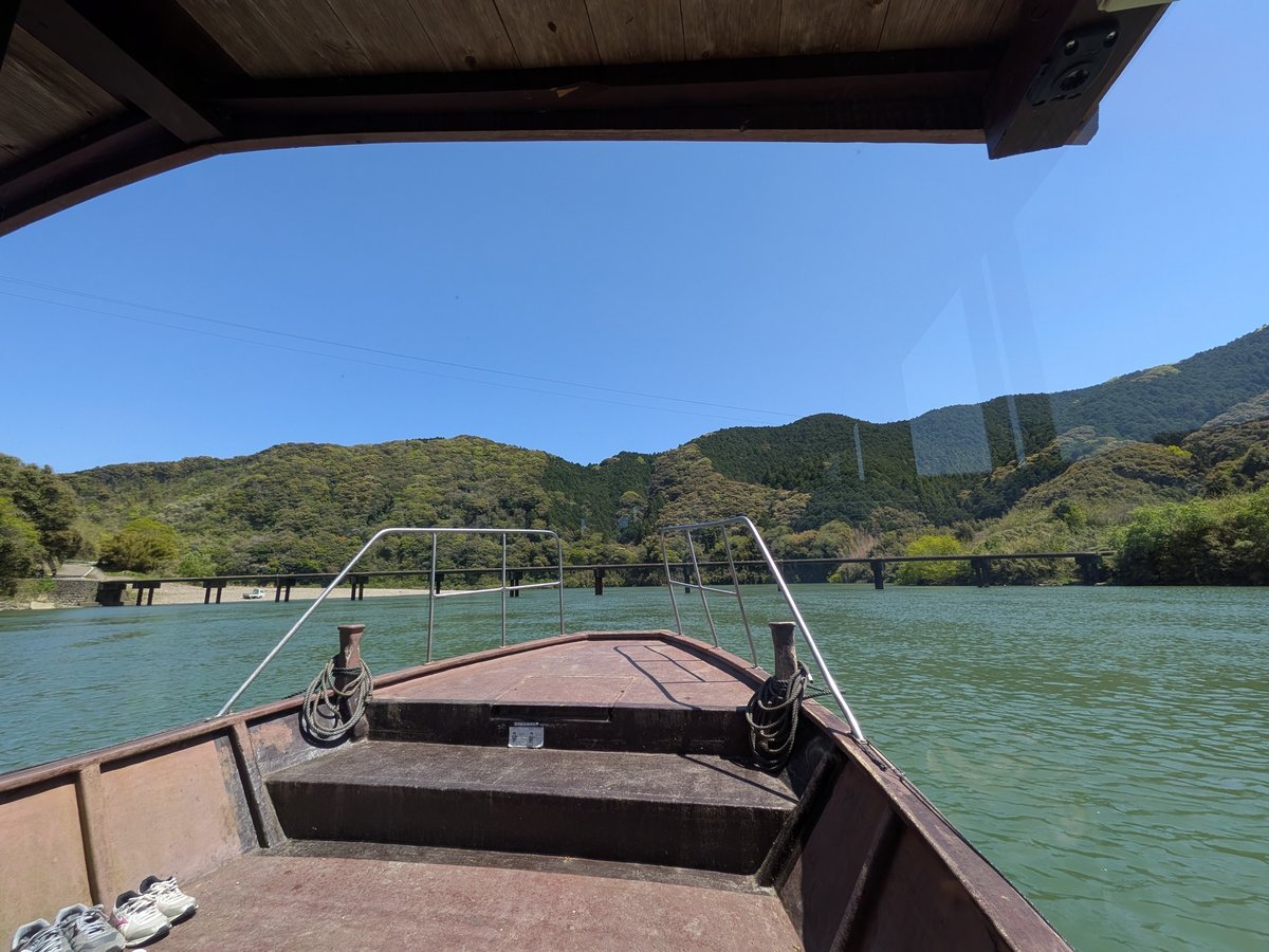 屋形船の船首から見た川と、遠くに見える橋と山々の景色。青い空が広がり、開放的な雰囲気が伝わる。