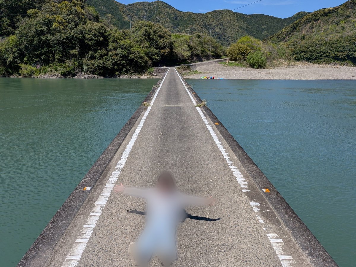四万十川に架かる沈下橋の上で、モザイク処理された人物が両腕を広げて寝そべっている様子。橋の向こうには川と緑豊かな山々が広がる。