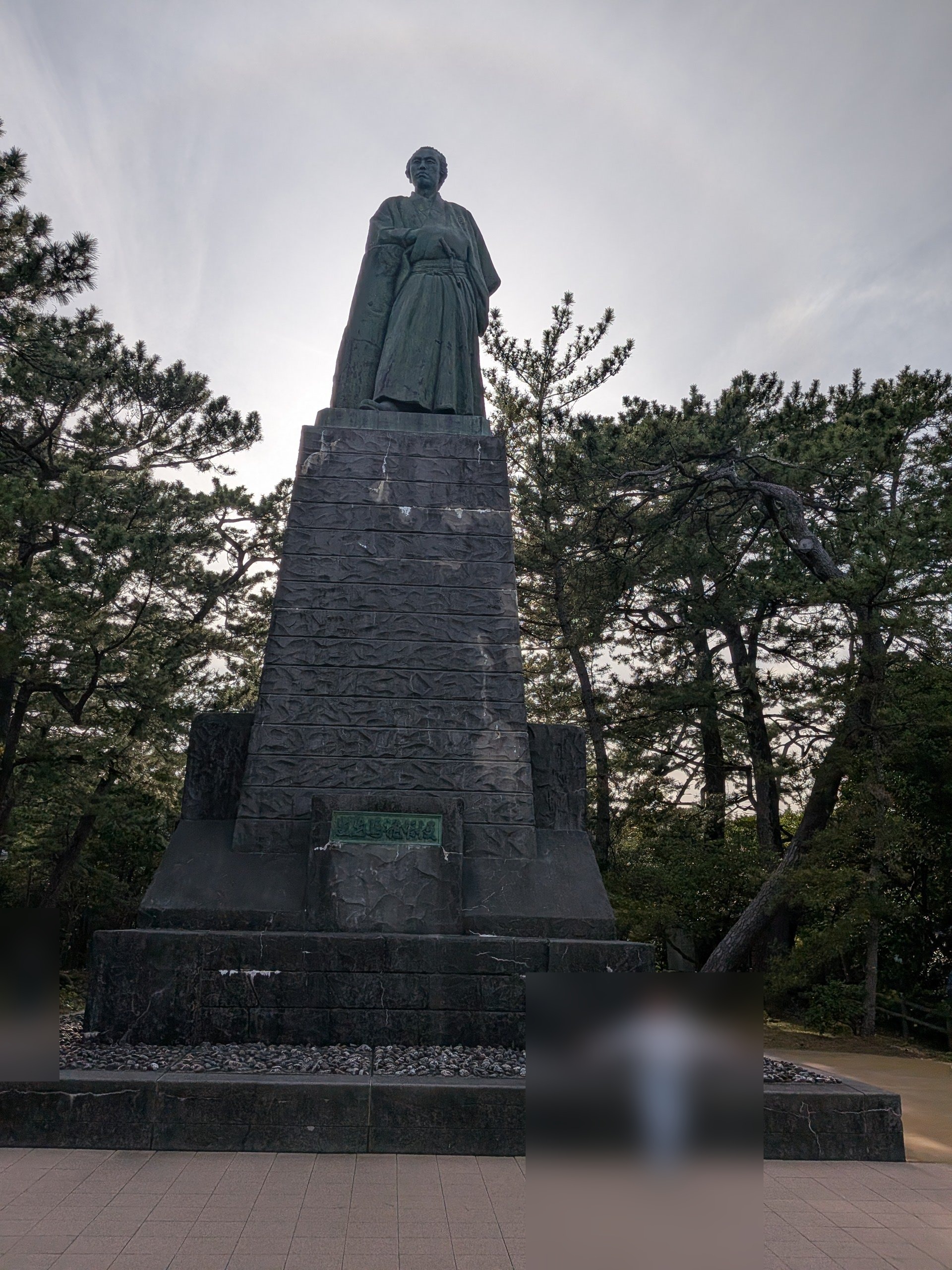 桂浜に立つ巨大な坂本龍馬像を下から見上げたアングル。像の周囲には松の木が茂り、歴史的な人物の存在感を際立たせている。